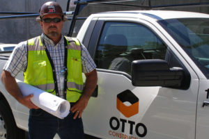 an image of an Otto worker standing in front of his truck in a green best and a hard hat on his head holding some construction blueprints
